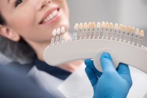 Blue gloved hand holding up an array of veneers with a woman smiling in the background